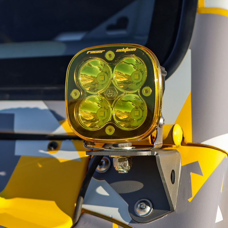 Baja Designs Squadron Single Rock Guard on a vehicle: an amber Squadron Racer Edition light on a pillar bracket of a yellow and gray camo wrapped vehicle, vehicle not identifiable