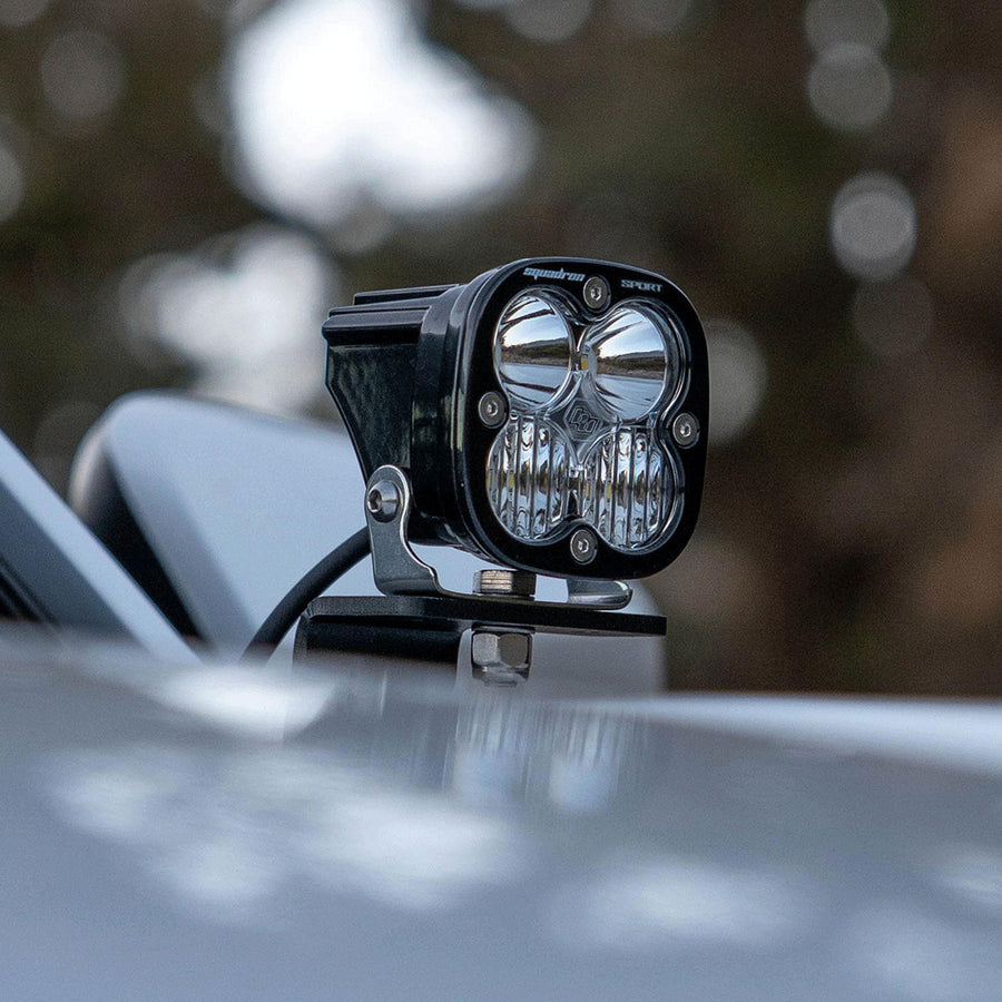 Baja Designs Squadron Sport A-Pillar Kit on a vehicle: A Squadron pod on an A-pillar bracket at the windshield base of a dark vehicle, trees blurred behind