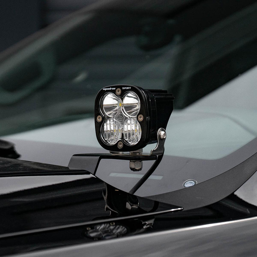 Baja Designs Squadron Sport A-Pillar Light Kit on a vehicle: A black Squadron Sport pod with a clear lens mounted on an A-pillar bracket beside the hood of a dark truck