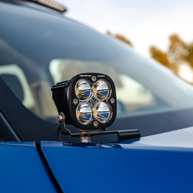 Baja Designs Squadron Sport A-Pillar Light Kit on a vehicle: A Squadron Sport LED pod with a clear lens on an A-pillar bracket at the base of a blue vehicle's windshield