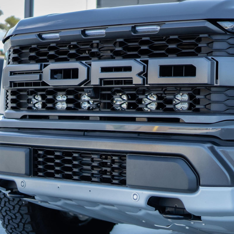 Baja Designs Squadron Sport Behind Grille Light Kit on a Ford F-150 Raptor: gray Ford Raptor grille with six square LED pods lit behind the lower grille below the FORD lettering