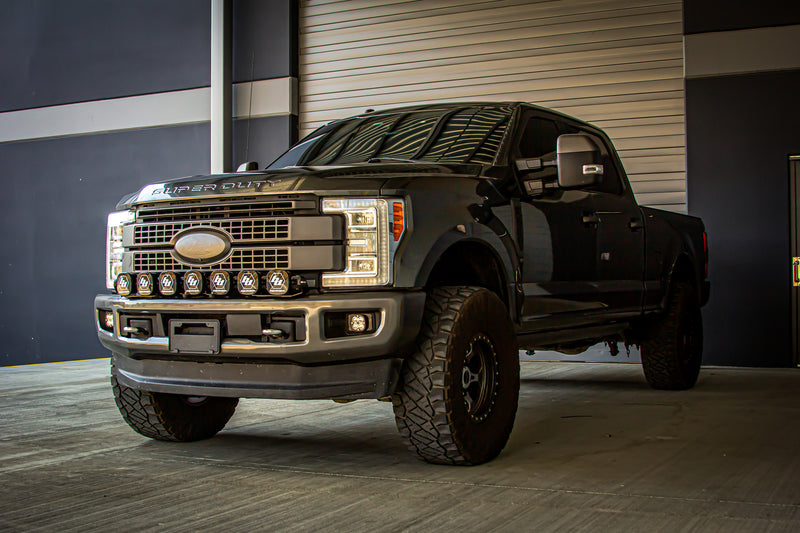 Baja Designs XL Linkable Bumper Light Kit installed on a Ford F-250 Super Duty: A dark gray Ford Super Duty pickup parked by a garage door with a seven-pod XL Linkable light bar mounted across the front bumper