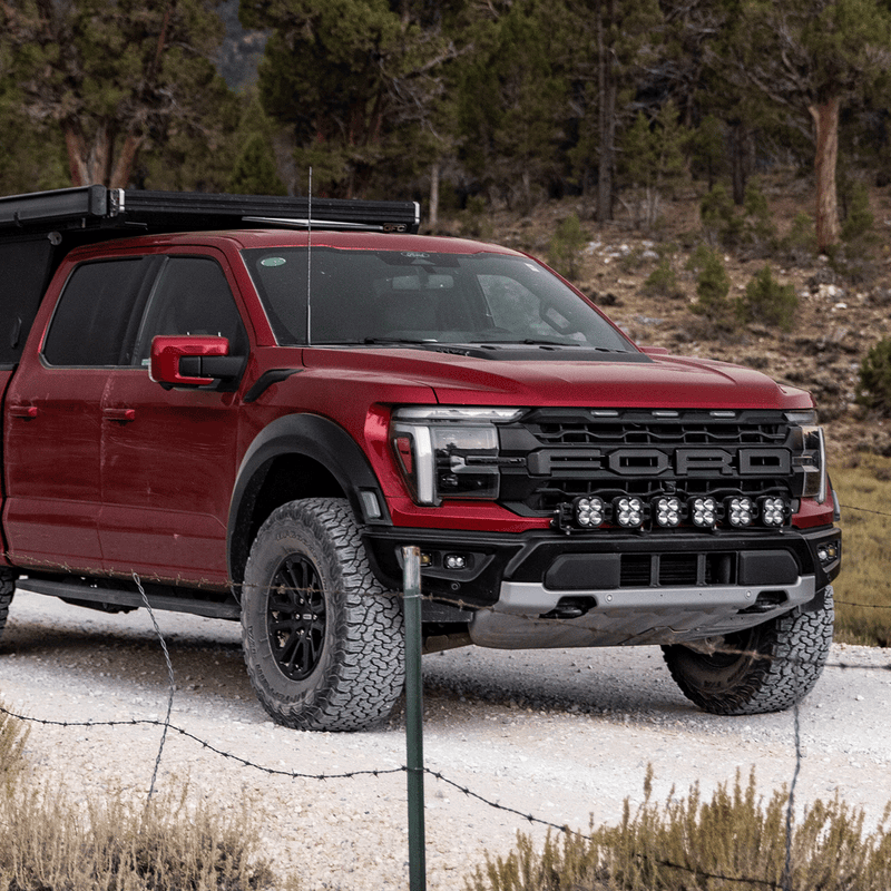Baja Designs XL Linkable Bumper Light Kit installed on a Ford F-150 Raptor: A red late-model Ford Raptor with a roof rack parked on a gravel road in the pines, with six XL pods under the grille
