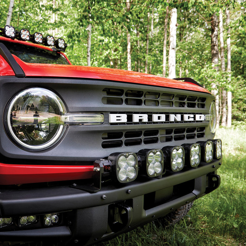 Baja Designs XL Linkable Bumper Light Kit on a Ford Bronco: front corner view of a red Ford Bronco with six XL linkable pods mounted across the front bumper in a forest