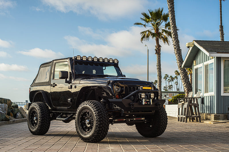 Baja Designs XL Linkable Roof Mount Light Kit installed on a Jeep Wrangler JK: A black two-door Jeep Wrangler with a linkable LED pod bar over the windshield is parked on a brick boardwalk by palm trees