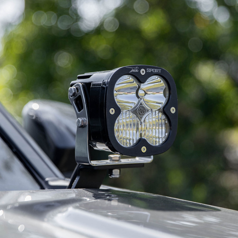 Baja Designs XL Sport A-Pillar Kit on a vehicle: A Baja Designs XL Sport LED pod mounted on an A-pillar bracket of a gray truck with trees behind it