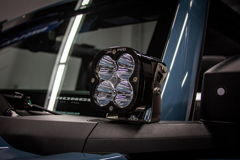 Baja Designs XL Sport A-Pillar Light Kit on a Ford Bronco: A black XL Pro pod with a clear lens mounted at the A-pillar of a dark blue Ford Bronco with door lettering visible