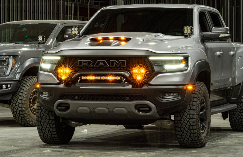 Baja Designs XL Sport A-Pillar Light Kit installed on a Ram 1500 TRX: A gray Ram TRX in a garage with A-pillar pods, glowing amber grille lights and a light bar, with a second truck parked behind
