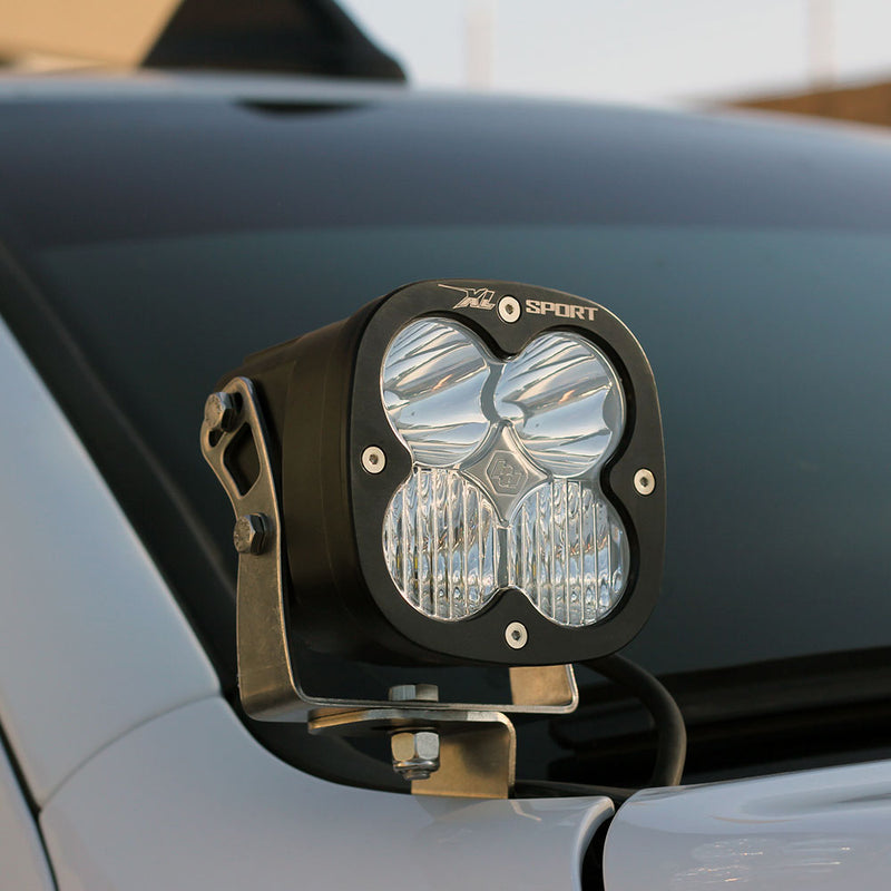 Baja Designs XL Sport LED Auxiliary Light Pod Pair on a vehicle: A black Baja Designs XL Sport LED pod with a clear lens mounted on a bracket beside the hood of a gray vehicle
