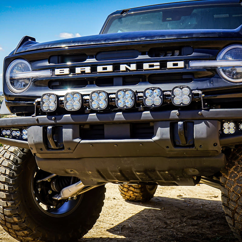 Baja Designs XL Sport Linkable LED Light Bar installed on a Ford Bronco: A dark blue Ford Bronco with a six-light XL Linkable LED bar mounted across its front bumper, parked on dirt under a blue sky