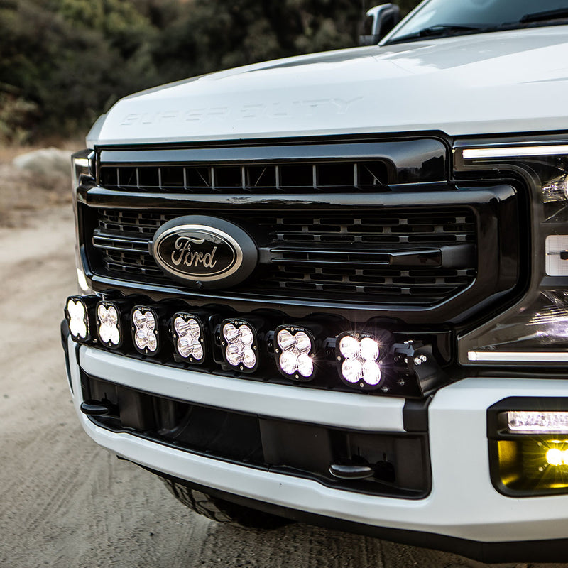 Baja Designs XL Sport Linkable LED Light Bar installed on a Ford Super Duty: A white Ford Super Duty pickup with a lit seven-light XL Linkable LED bar mounted in its front bumper on a dirt road