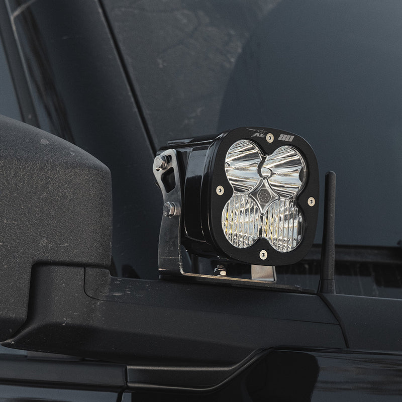 Baja Designs XL80 A-Pillar Light Kit on a vehicle: A black XL 80 pod with a clear lens mounted on an A-pillar bracket beside the windshield of a dark vehicle