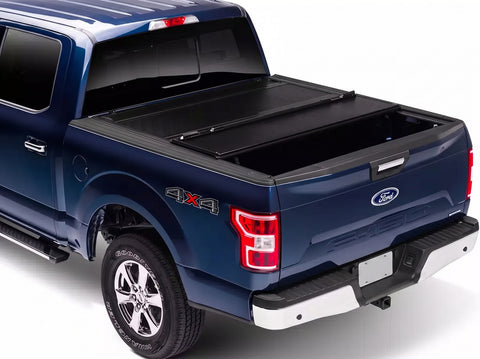 BAKFlip FiberMax hard folding tonneau cover in matte black folded forward against the cab of a blue Ford F-150 pickup, shown from the rear passenger side with the bed open