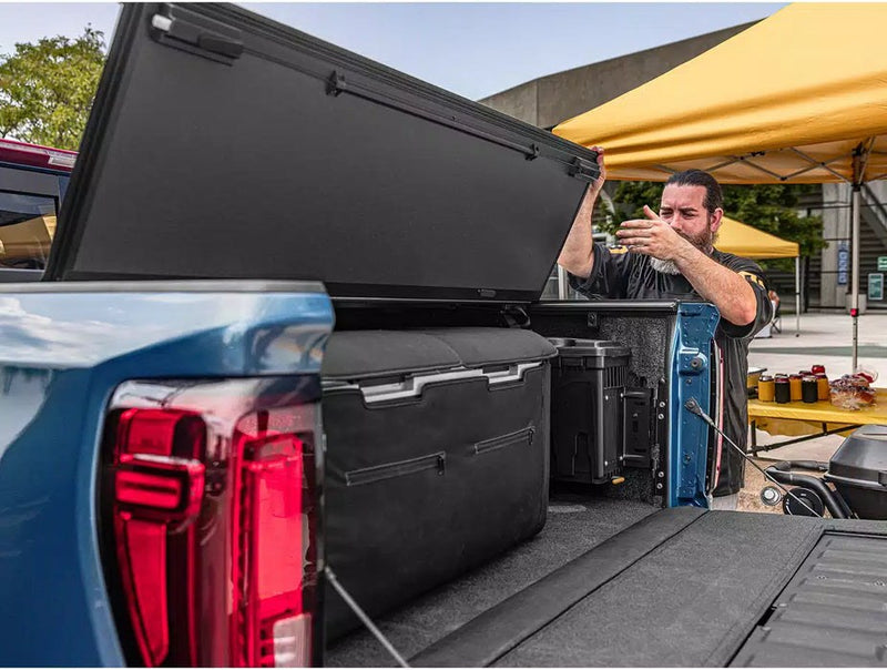 Man folding the black BAKFlip MX4 hard tonneau cover panels upward on a blue pickup with the tailgate down and storage cases in the bed at an outdoor tailgate event with a yellow canopy