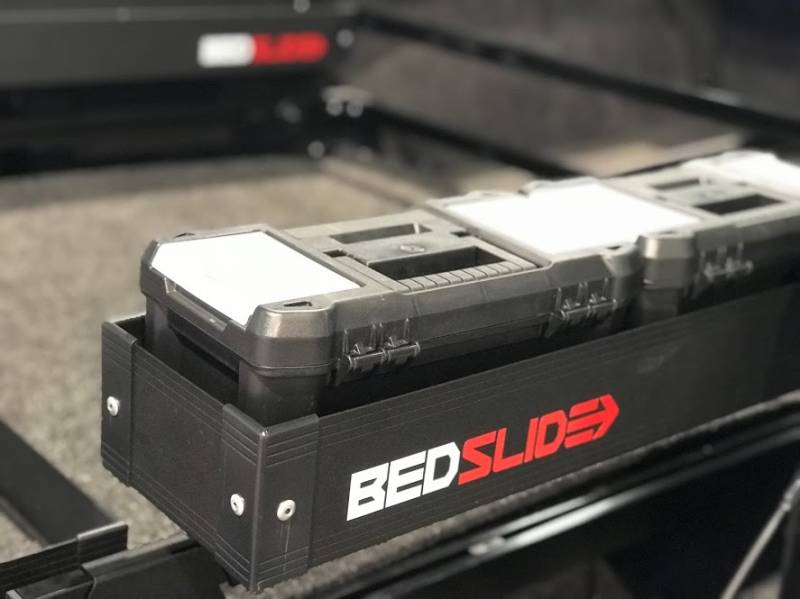 Close-up of a black BEDSLIDE BEDBIN loaded with stacked tool cases, showing the white BEDSLIDE decal on the bin