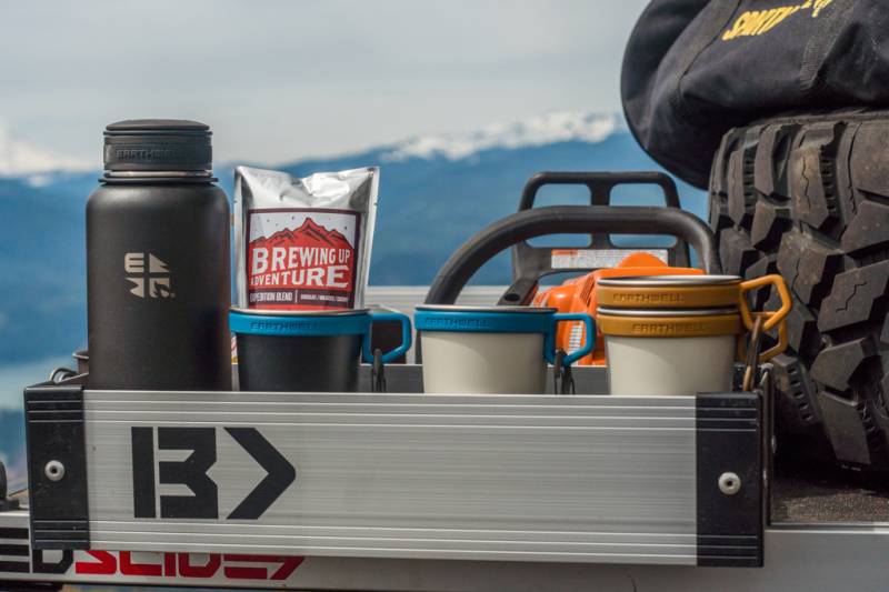 Silver Mini Kix bin holding a flask, mugs and camp supplies on a tailgate at a mountain overlook