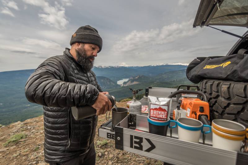 Person making coffee at the open tailgate of a truck with the silver Mini Kix bin holding the supplies