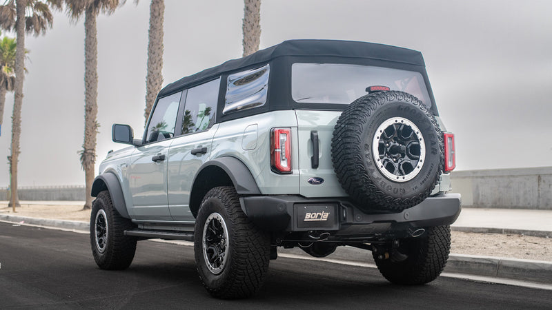 Borla ATAK Axle-Back Exhaust System installed on a Ford Bronco 4-door: A light gray four-door Ford Bronco with a soft top parked by palm trees seen from the rear quarter