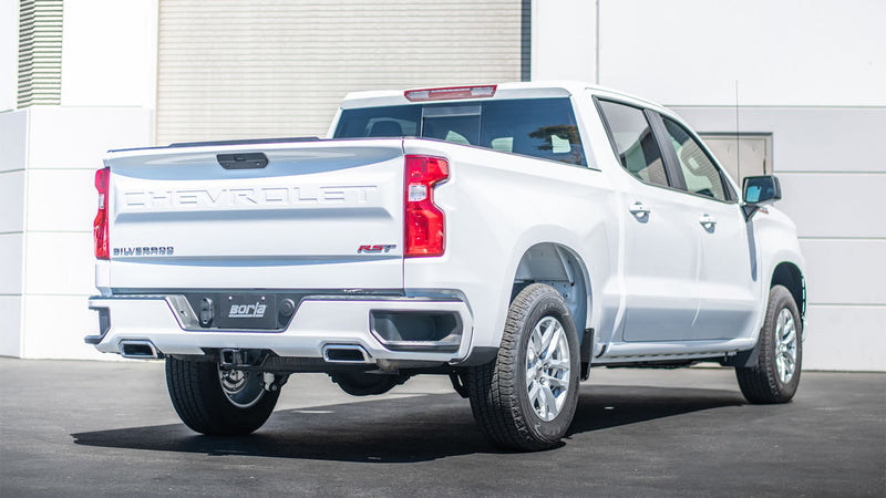 Borla Touring Cat-Back Exhaust System installed on a Chevrolet Silverado 1500: A white Chevrolet Silverado 1500 crew cab parked in front of a building, rear view showing exhaust tips exiting on both sides of the rear bumper