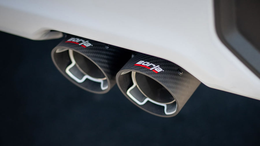 Cat-Back Exhaust Systems by Borla: A close-up of twin carbon fiber Borla exhaust tips exiting beneath a white rear bumper.