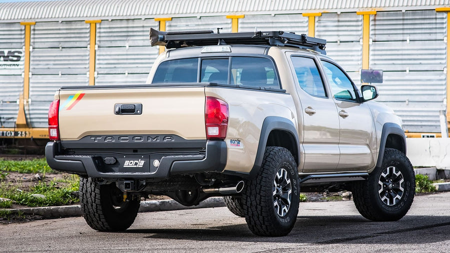 Cat-Back Exhaust Systems by Borla: Tan Toyota Tacoma with roof rack seen from the rear quarter showing a polished side-exit tip ahead of the rear wheel and a Borla plate, freight railcars behind.
