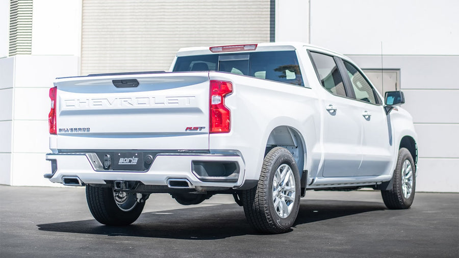 Cat-Back Exhaust Systems by Borla: A white Chevrolet Silverado 1500 crew cab parked in front of a building, rear view showing exhaust tips exiting on both sides of the rear bumper.