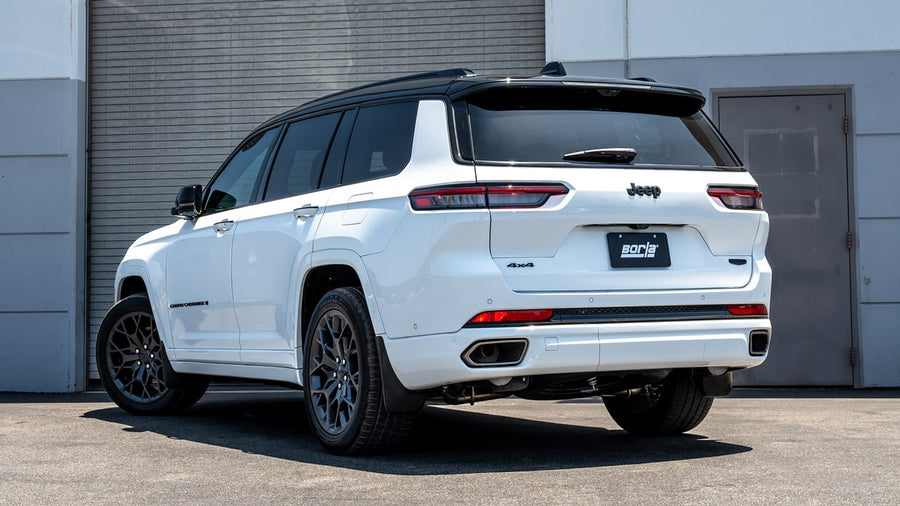 Cat-Back Exhaust Systems by Borla: White Jeep Grand Cherokee L 4x4 from the rear quarter in front of a roll-up door with a Borla plate.