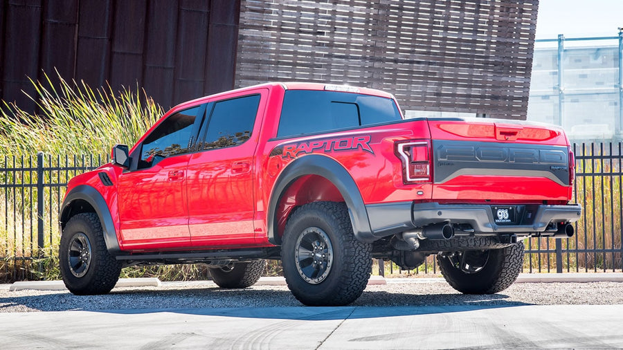 Mid-Section Exhaust Systems by Borla: Red Ford F-150 Raptor parked by a fence, rear three-quarter view showing polished exhaust tips exiting ahead of the rear tires.