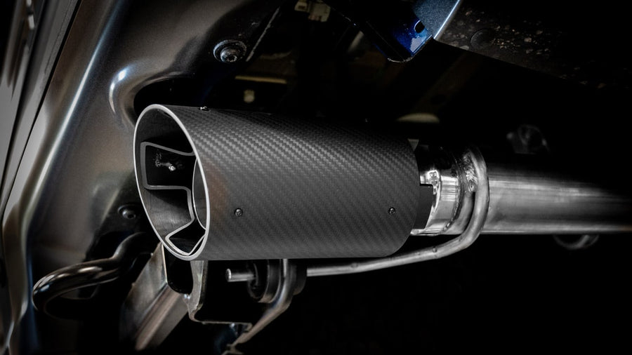 Tail Pipe & Connecting Pipe Kits by Borla: A carbon fiber exhaust tip is shown up close mounted under a vehicle against a dark background.