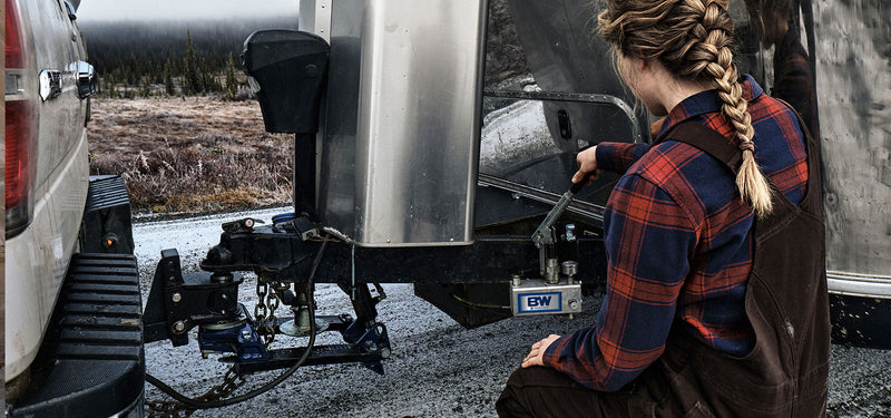 Person kneeling beside a coupled travel trailer, pumping the hydraulic handle on a B&W Continuum weight distribution hitch on a gravel pull-off