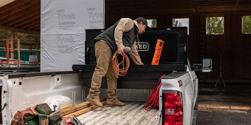 Contractor loading gear into a black DECKED Tool Box mounted in a white pickup bed on a framing jobsite