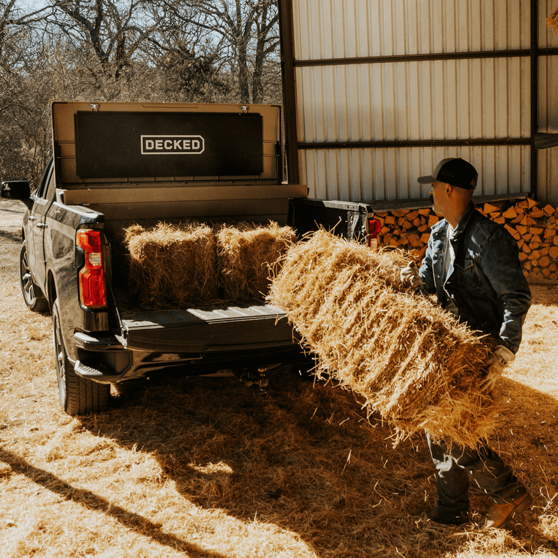 Fender Dirt tan DECKED Tool Box open in the bed of a black pickup while a rancher loads hay bales behind it