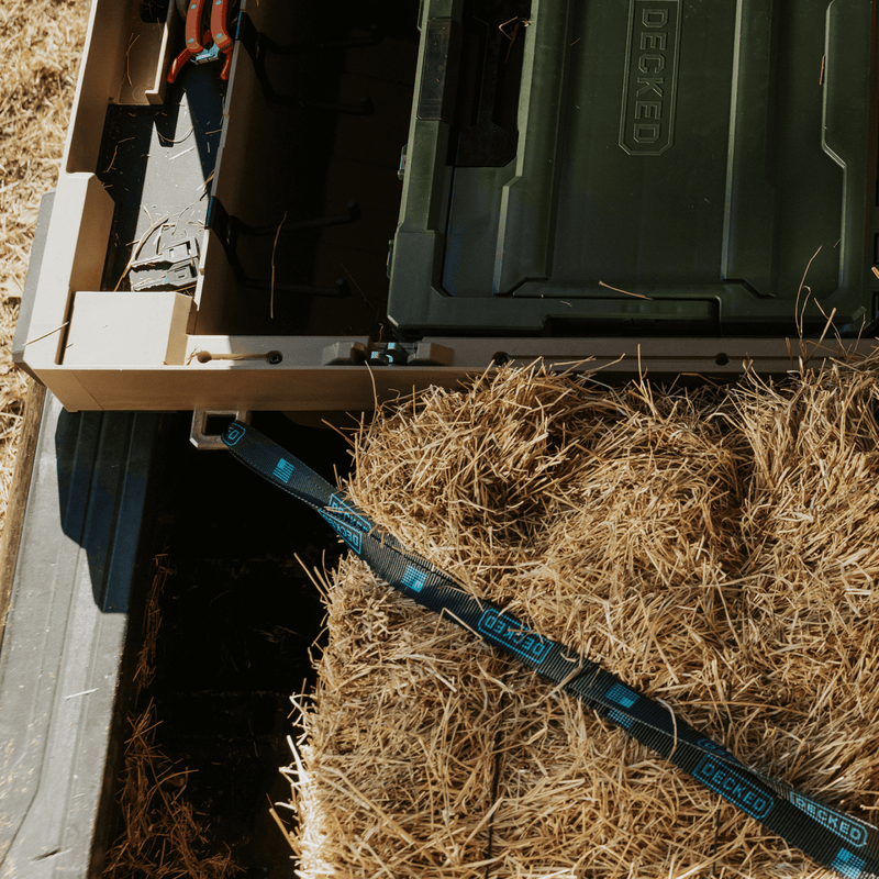 Close-up of a DECKED ratchet strap anchored below a Fender Dirt tan DECKED Tool Box while the bed below carries hay bales