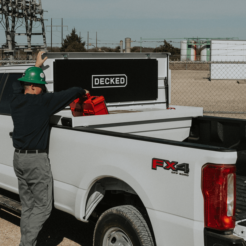 Worker in a green hard hat reaching into an open Industrial White DECKED Tool Box in a white pickup bed at a utility substation