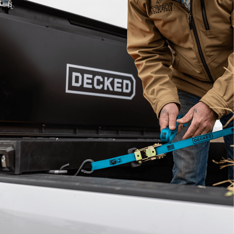 Hands tightening a DECKED ratchet strap on the tie-down point below a closed black DECKED Tool Box
