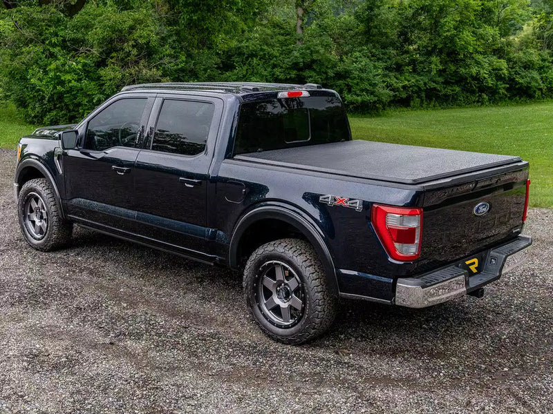 Soft tri-fold cover closed on a dark pickup parked on gravel by trees