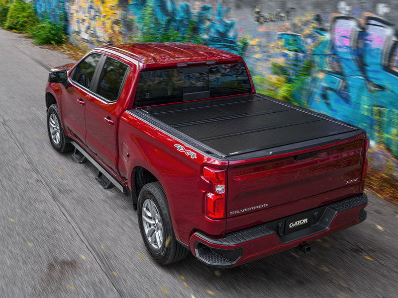 Gator FX tonneau cover (closed) on a Chevrolet Silverado 1500: A red Chevrolet Silverado with a closed black hard folding tonneau cover drives past a graffiti wall