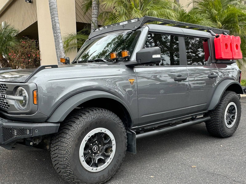 Go Rhino 4000 Series: gray 4-door Bronco parked by palm trees with black round side steps below the doors, plus a roof rack and red fuel cans (shown with roof rack, auxiliary pod lights, fuel cans, aftermarket front bumper, not included)