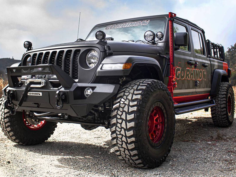 Go Rhino Exterior Jack Mount: lifted black and red Jeep Gladiator on gravel with a red farm jack mounted at the windshield pillar (shown with Hi-Lift style farm jack, front bumper, winch, lights, side steps, bed rack, lift kit, aftermarket wheels and