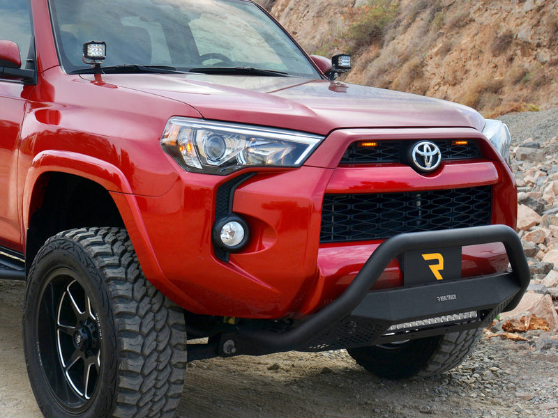 Go Rhino RC4 LR: red SUV front corner on a rocky trail with a textured black bull bar and LED light bar (shown with LED light bar, A-pillar cube lights, aftermarket wheels and tires, not included)