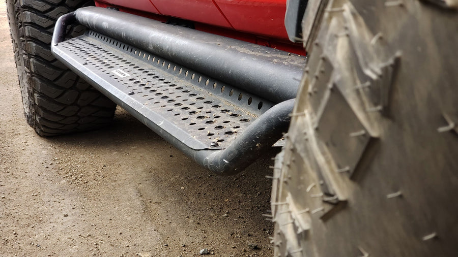 Go Rhino Dominator D1 Side Steps: Black tube side step with perforated step plate installed under a red vehicle between large off-road tires