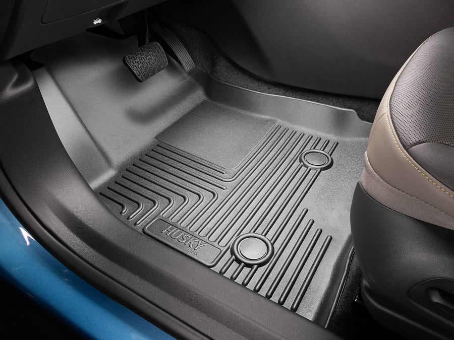 Husky Liners X-act Contour Floor Liners: Black Husky floor liner installed in a driver footwell beside a gray leather seat, pedals above, blue door sill edge visible
