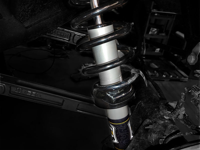 ICON Vehicle Dynamics 2.5 EXP Series Coilover on a vehicle: an installed ICON 2.5 coilover with black spring and silver body inside a wheel well, ICON-embossed control arm behind, in a shop