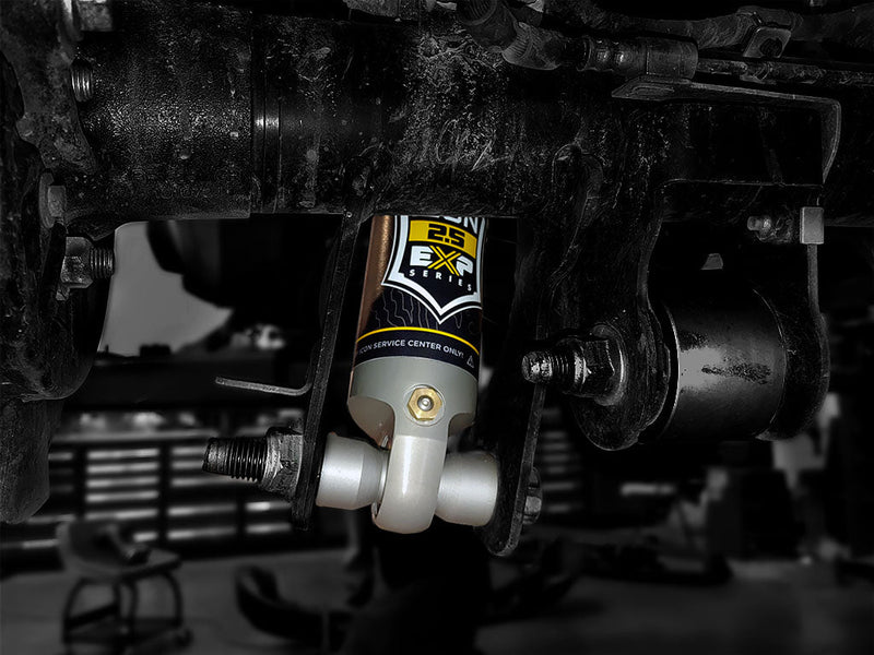 ICON Vehicle Dynamics 2.5 EXP Series Coilover on a vehicle: close view of the lower end of an installed ICON 2.5 EXP shock bolted into an axle bracket under a dirty vehicle in a shop (axle and bracket shown, not included)