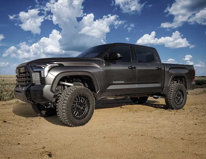 A gray third-generation Toyota Tundra crew cab with lift and off-road tires parked on sand under a cloudy sky.