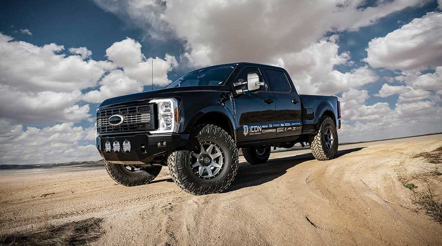 A black lifted Ford Super Duty crew cab with aftermarket front bumper, light pods and off-road tires on a sandy track under clouds.
