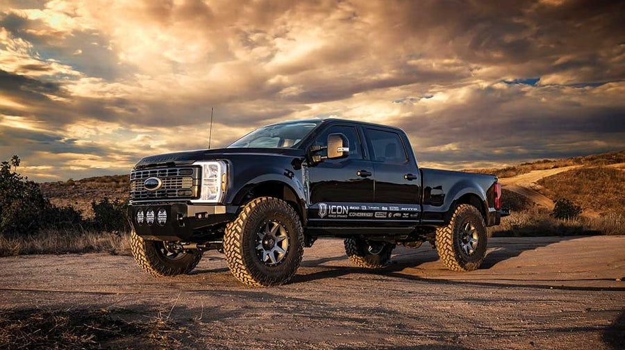 A black lifted Ford Super Duty crew cab with aftermarket bumper and light pods parked on dirt under a golden sunset sky.