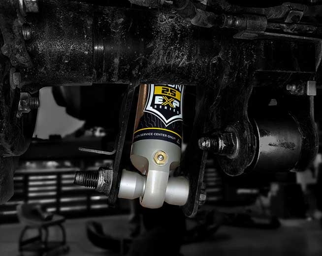 Close view of the lower end of an installed ICON 2.5 EXP shock bolted into an axle bracket under a dirty vehicle in a shop.