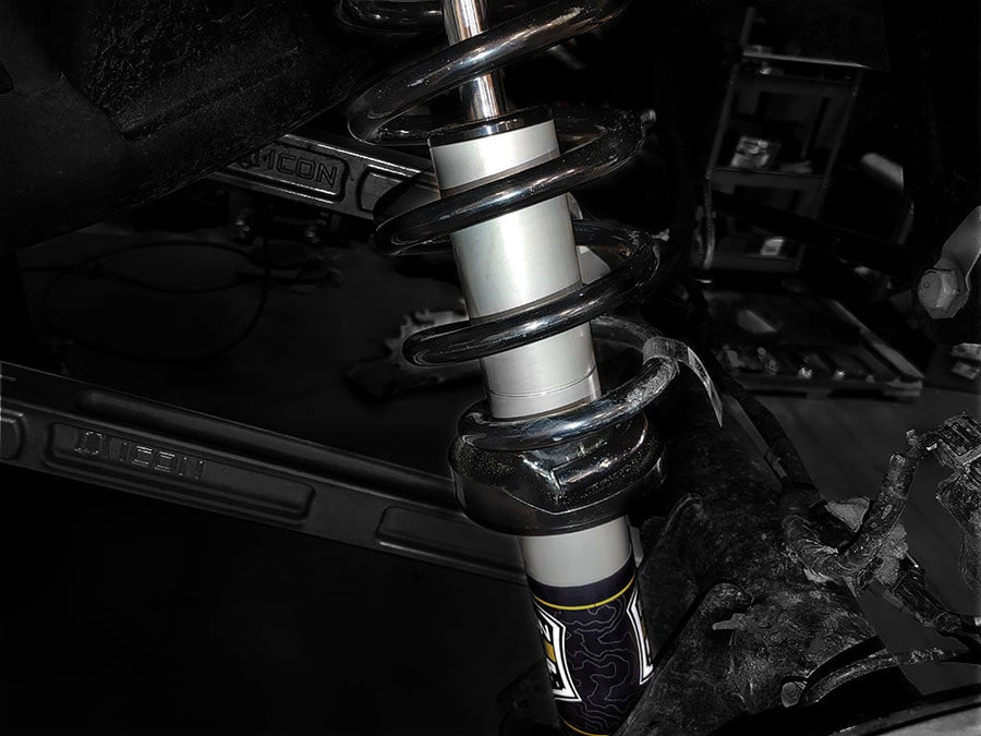 An installed ICON 2.5 coilover with black spring and silver body inside a wheel well, ICON-embossed control arm behind, in a shop.