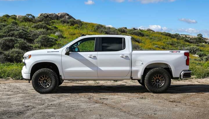 A white Chevrolet Silverado 1500 crew cab with bronze wheels and all-terrain tires in side profile on a dirt lot before a green hillside.